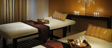 Ein Spa-Raum mit zwei Massagetischen, jeder Tisch mit Ablage und Kerze, sowie eine Vase mit weißen Blumen an der Wand.