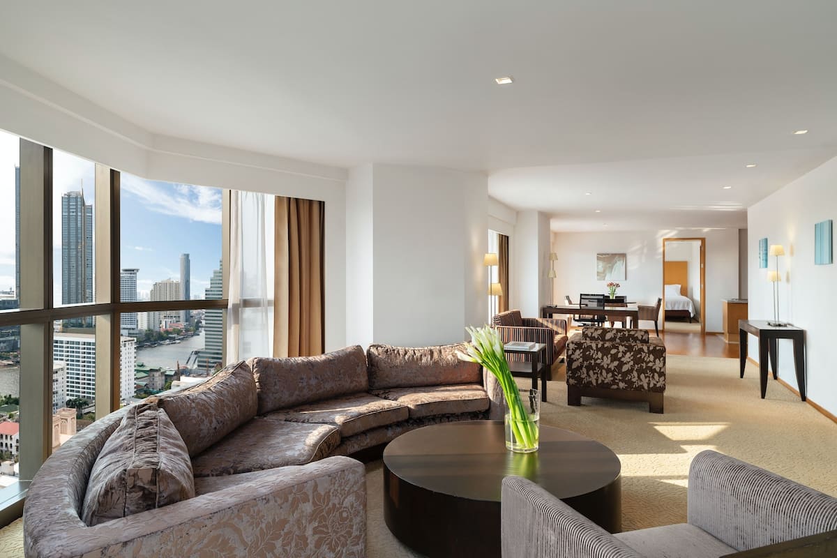 A modern hotel room with a large curved sofa, a round coffee table, and a view of the cityscape through large windows.