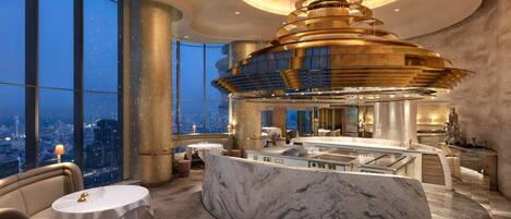 A luxurious hotel interior with a central marble bar, a high ceiling with a unique chandelier, and large windows offering a city view.