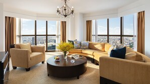 A spacious living room with a large sofa, armchairs, and a coffee table. A chandelier hangs from the ceiling, and the room features large windows with curtains.