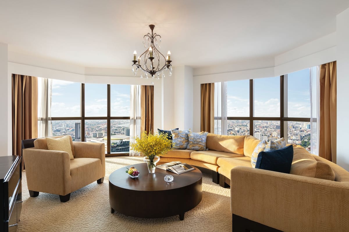 A spacious living room with a large sofa, armchairs, and a coffee table. A chandelier hangs from the ceiling, and the room features large windows with curtains.