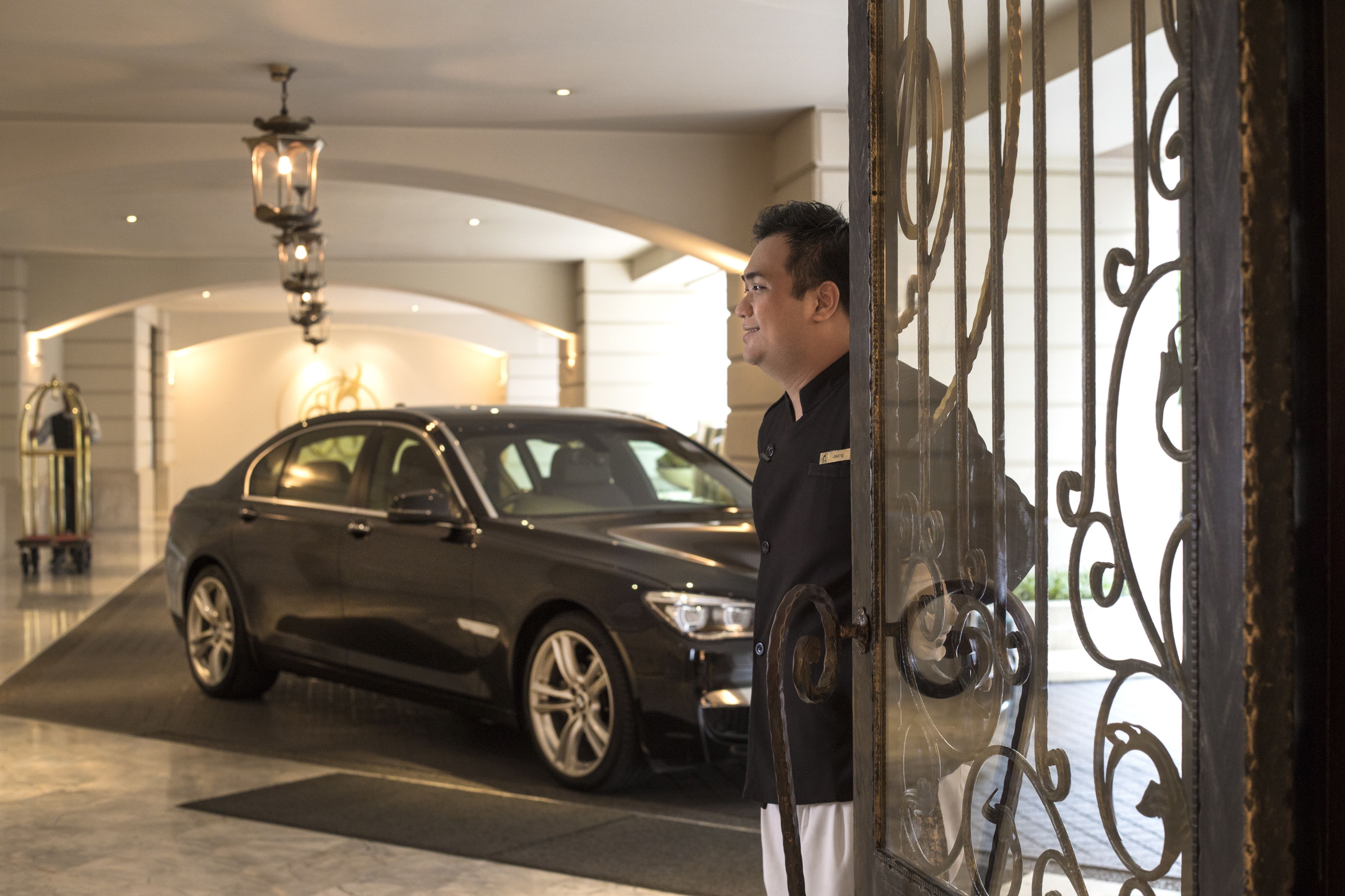 A man standing by a black car in a hotel lobby with a chandelier and a decorative iron gate.