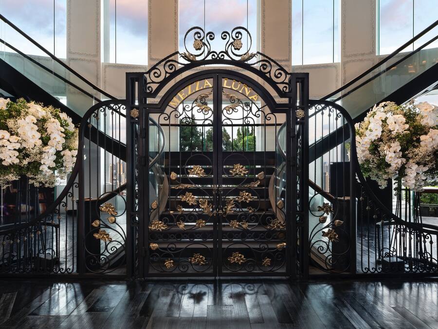 A grand entrance with a black wrought iron gate, white flowers, and the name "MEILA LUNA" above the stairs.
