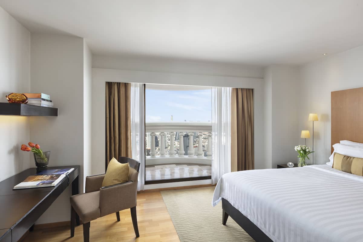 A hotel room with a bed, a desk, a chair, a view of the city through large windows, and a balcony with a railing.