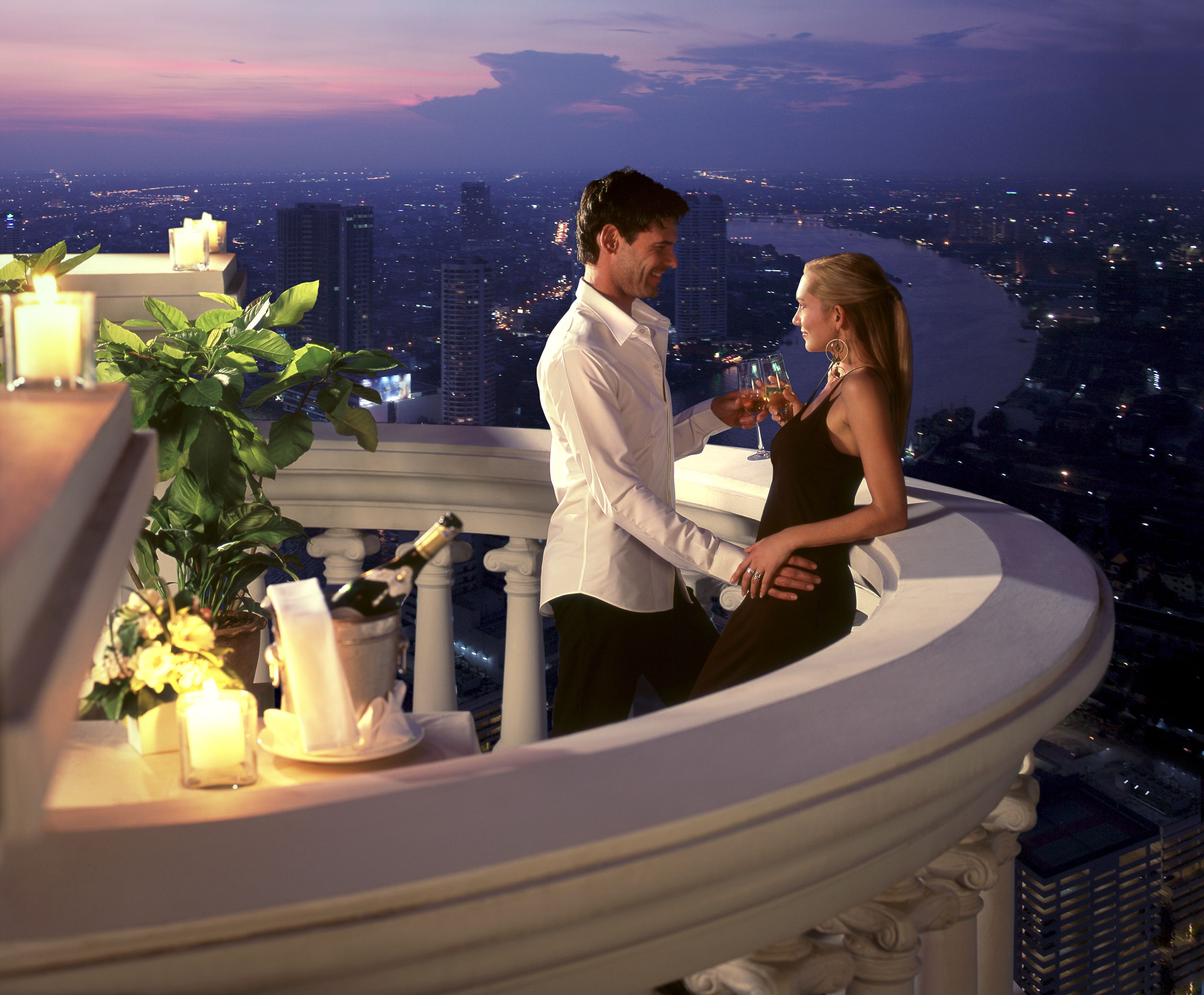 Um casal em uma varanda com vista para a cidade, uma mesa com velas e uma garrafa de champanhe.