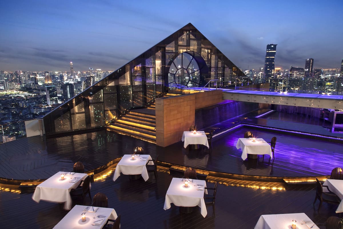A rooftop restaurant with tables set for dining, a glass structure, and a cityscape view.