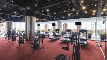Academia com diversos equipamentos de exercício, piso de carpete vermelho e grandes janelas com vista para edifícios e plantas lá fora.
