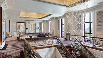 A grand hotel lobby with a spacious seating area, a high ceiling with ornate chandeliers, and a view of the city through large windows.