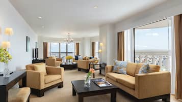 A spacious living room with beige sofas, a coffee table, and a TV. Large windows offer a view of the city and the sea.