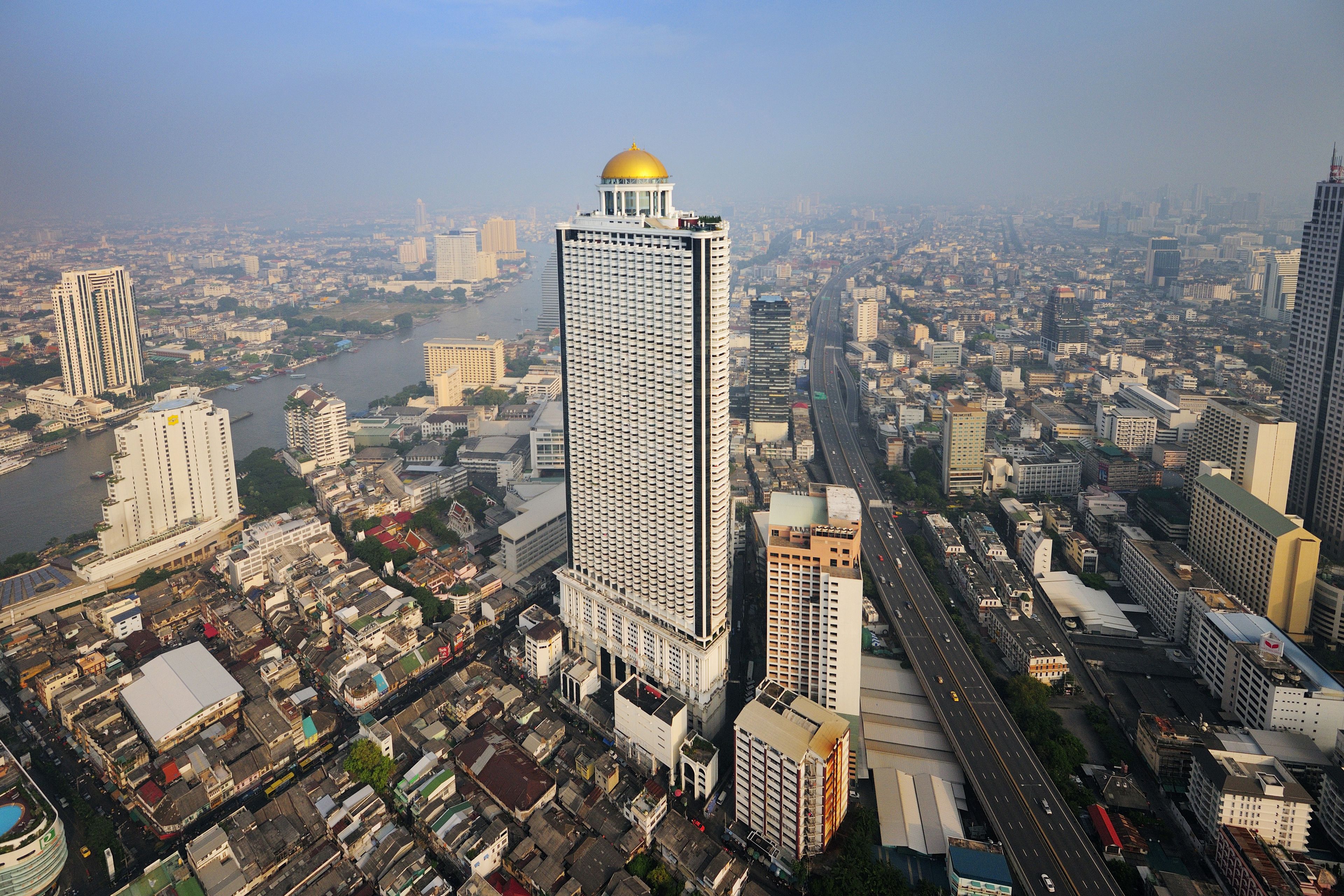 A high-rise building with a distinctive dome and a grid-like facade, surrounded by a dense urban environment with various other buildings and a river in the background.