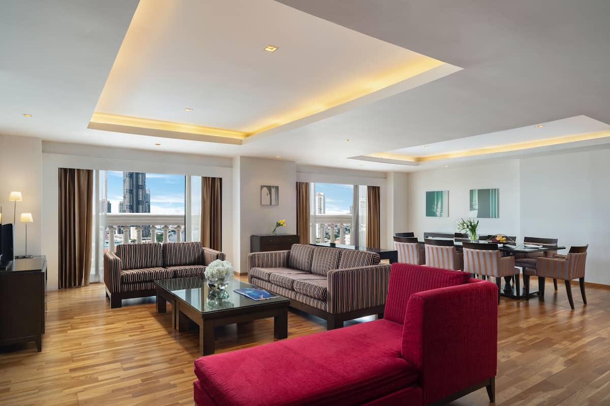 A spacious living room with a red sofa, striped couches, a dining area with chairs, and a view of the city through large windows.