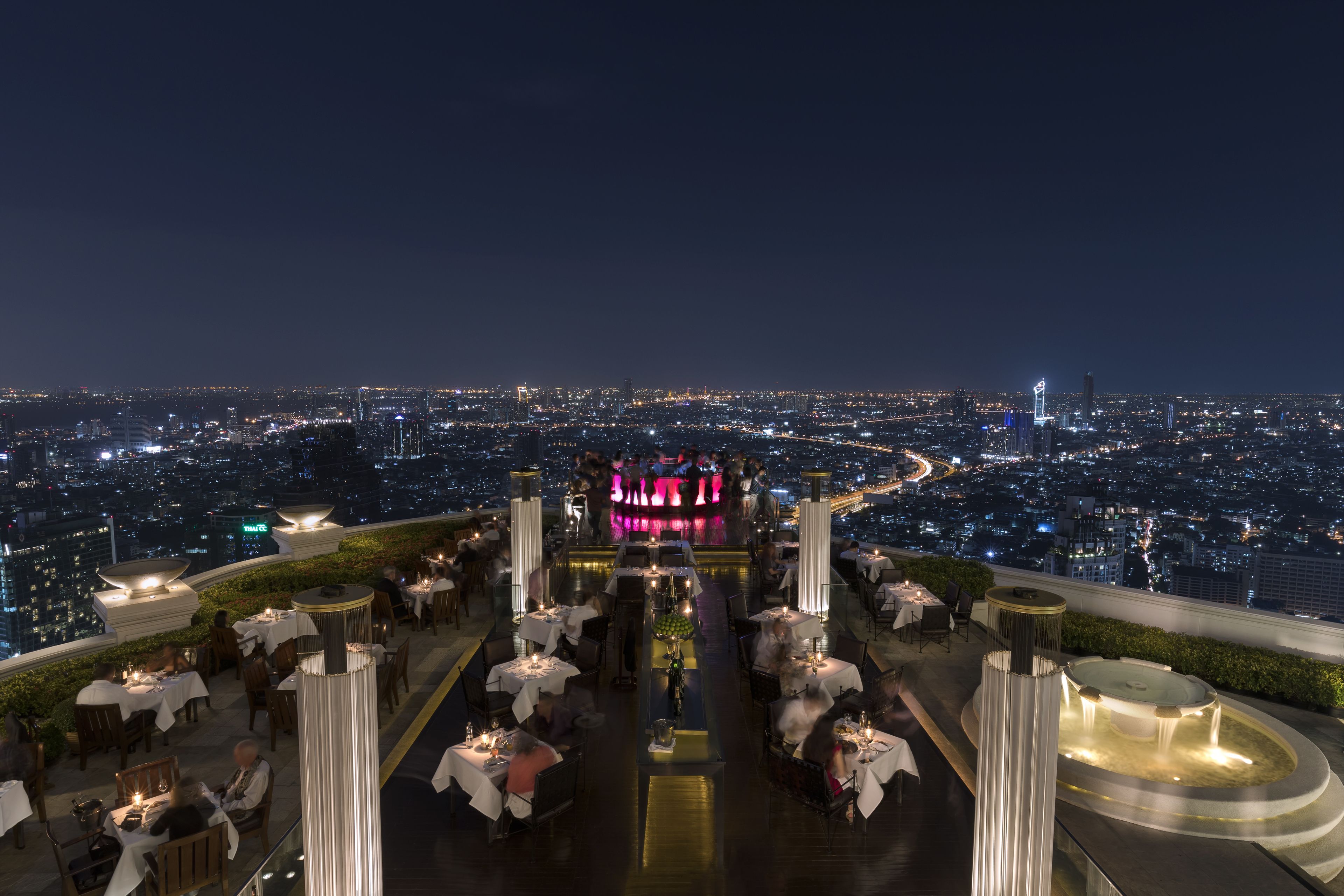 A rooftop restaurant with tables and chairs arranged around a central area with a fountain.