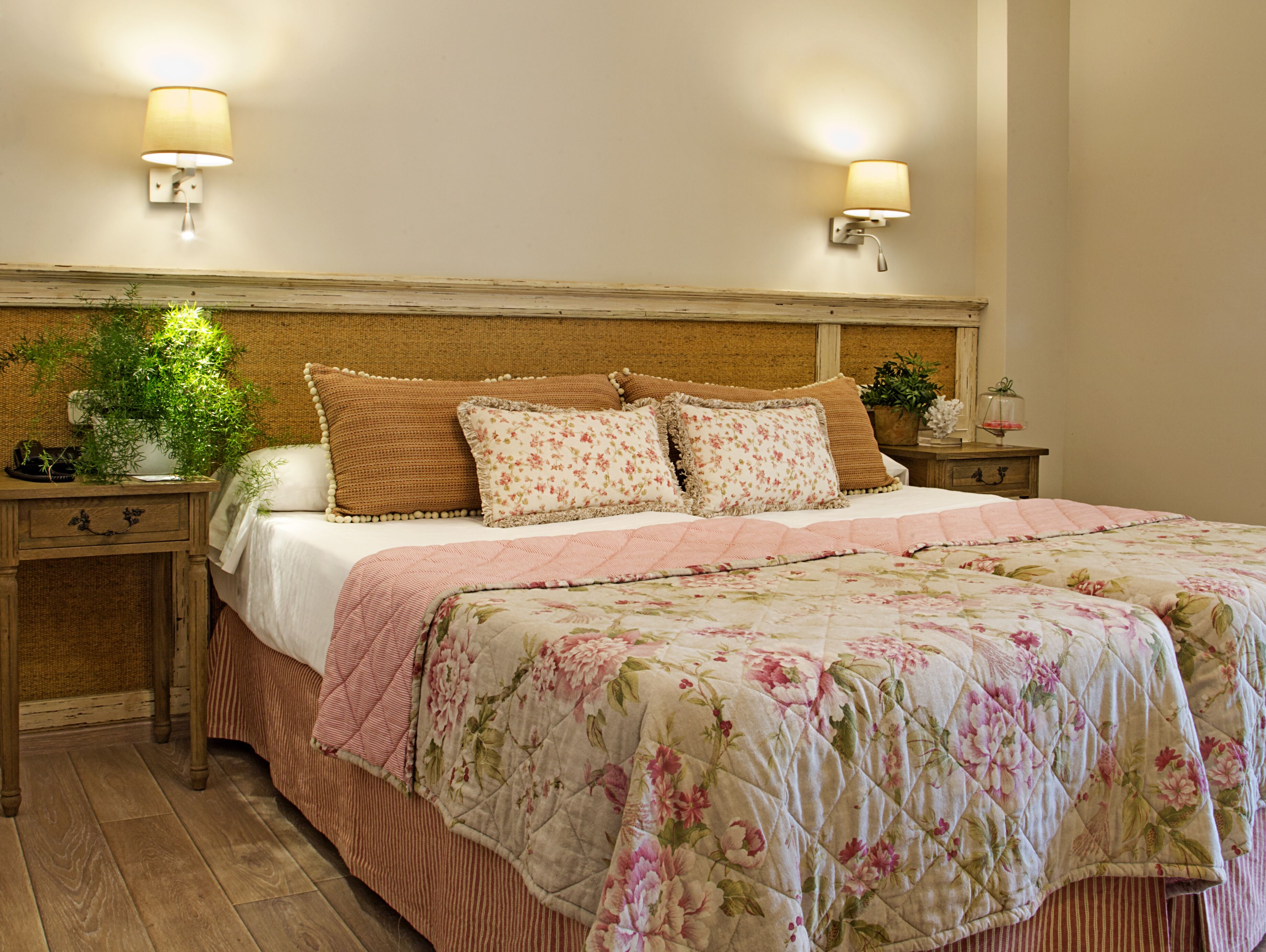 A bedroom with a wooden headboard, two bedside tables, a nightstand, a potted plant, and a floral bedspread.