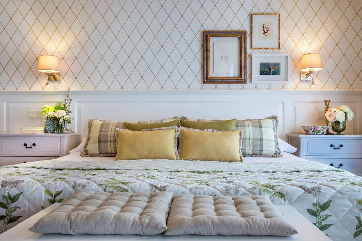 Una cama bien tendida con colcha floral, dos mesitas de noche con lámparas, y una pared con patrón geométrico y cuadros enmarcados.