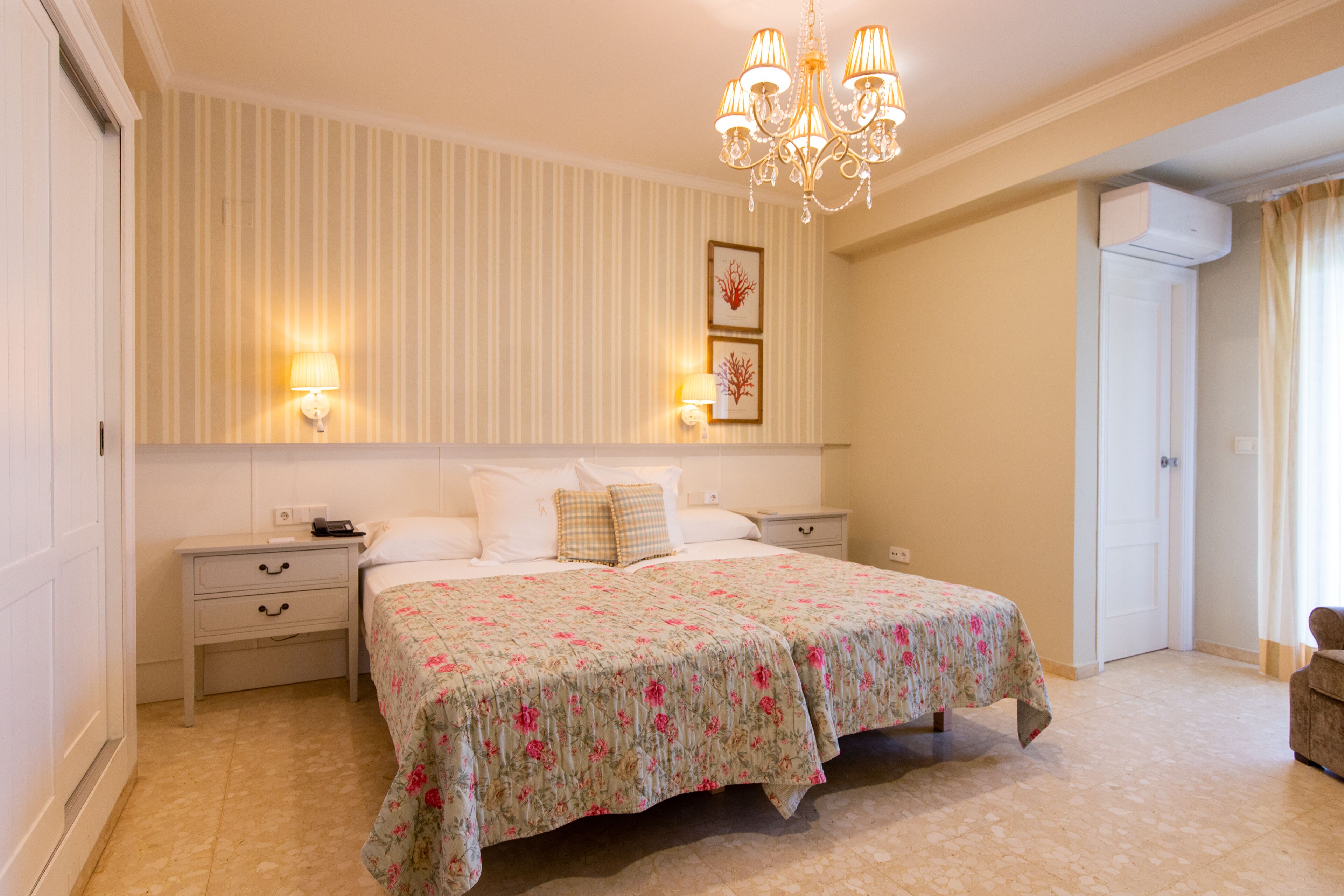 A bedroom with a large bed, two bedside tables, a chandelier, and a wall-mounted air conditioner.