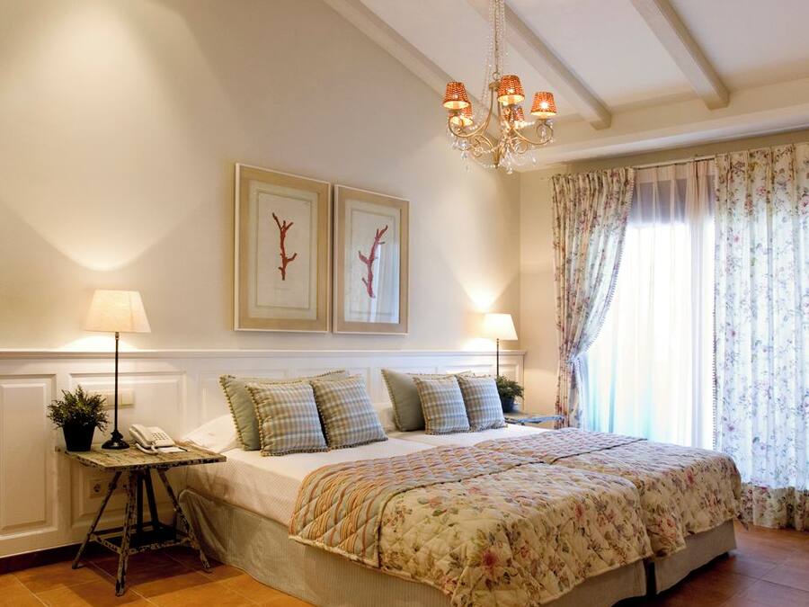 Un dormitorio con una cama grande, una araña, una mesita de noche con lámpara y dos cuadros en la pared.