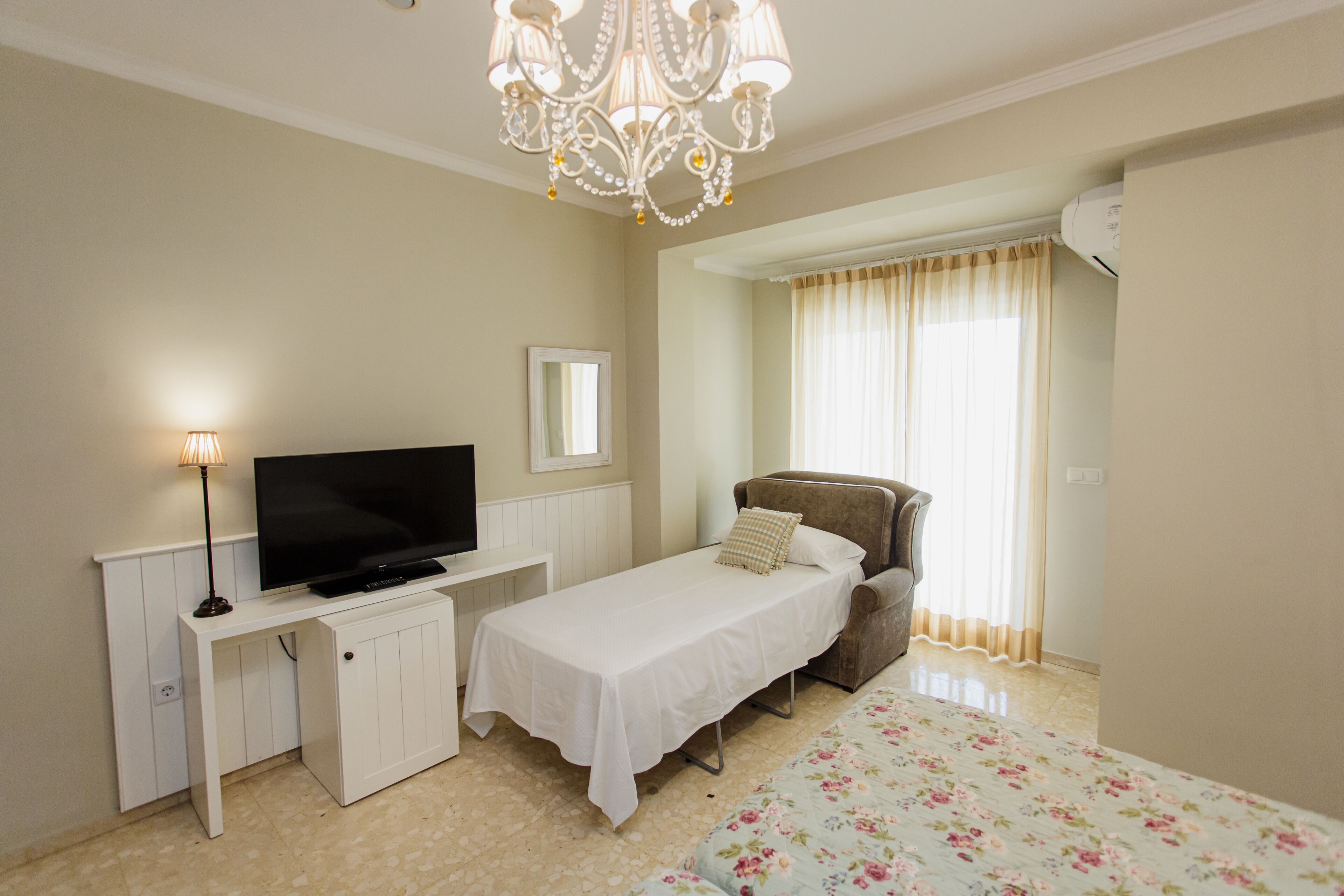 A bedroom with a bed, a TV, a desk, a chair, a chandelier, and a window with curtains.