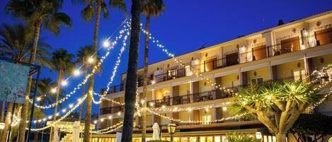 Un hotel con varios balcones, luces de adorno, palmeras y mobiliario exterior.