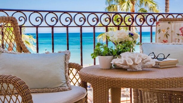 A balcony with wicker furniture, a potted plant, and a book. - Hotel Los Ángeles (Denia)
