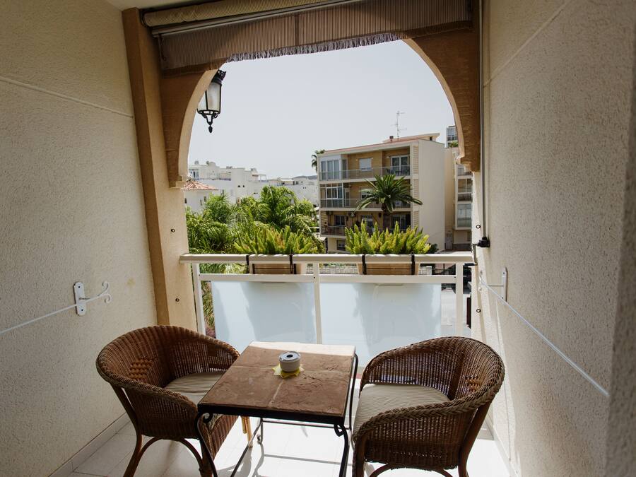 A balcony with wicker chairs and a table, overlooking a residential area with buildings and greenery.