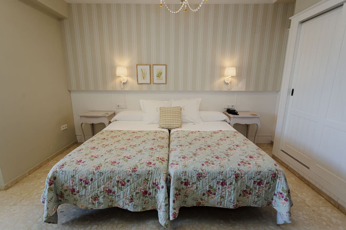 Cama doble con ropa de cama floral, dos mesitas de noche con lámparas, un candelabro y una puerta a la derecha.