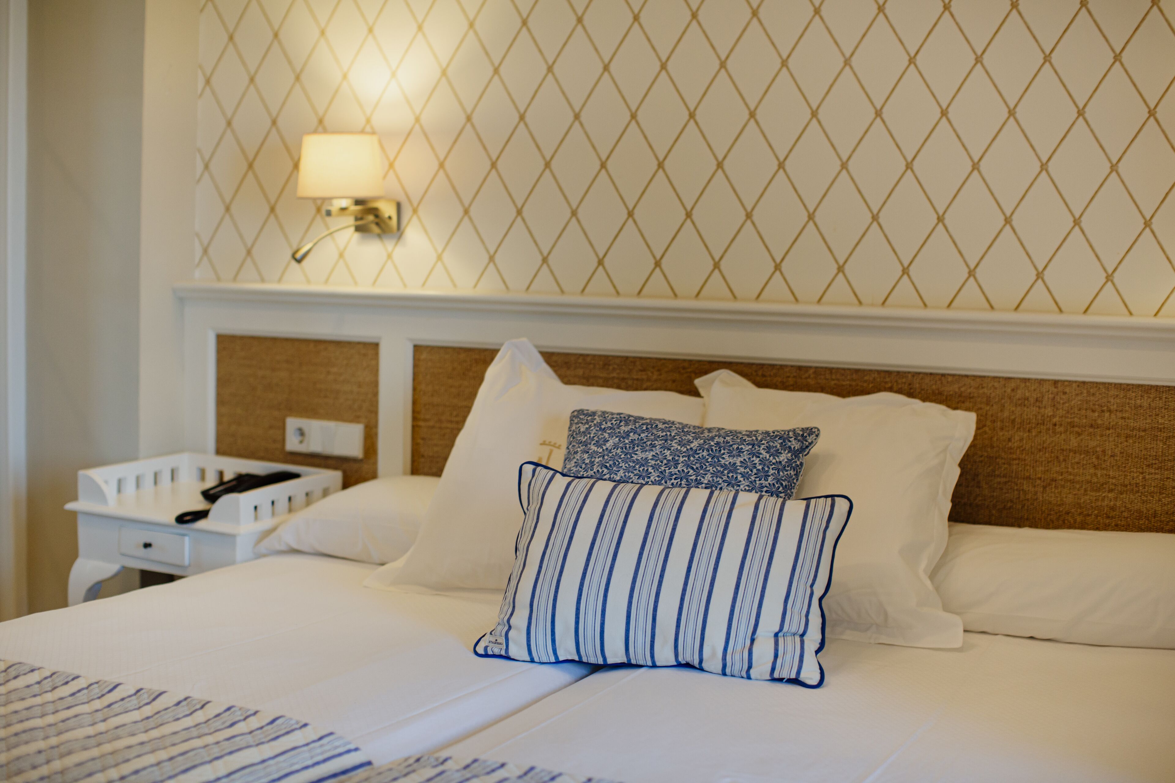 A neatly made bed with white linens and blue-striped pillows, a bedside table with a lamp, and a wall with a geometric pattern.