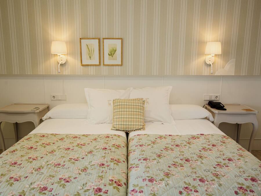 Habitación de hotel con una cama grande, dos mesitas de noche, una lámpara de pared e impresiones botánicas enmarcadas.