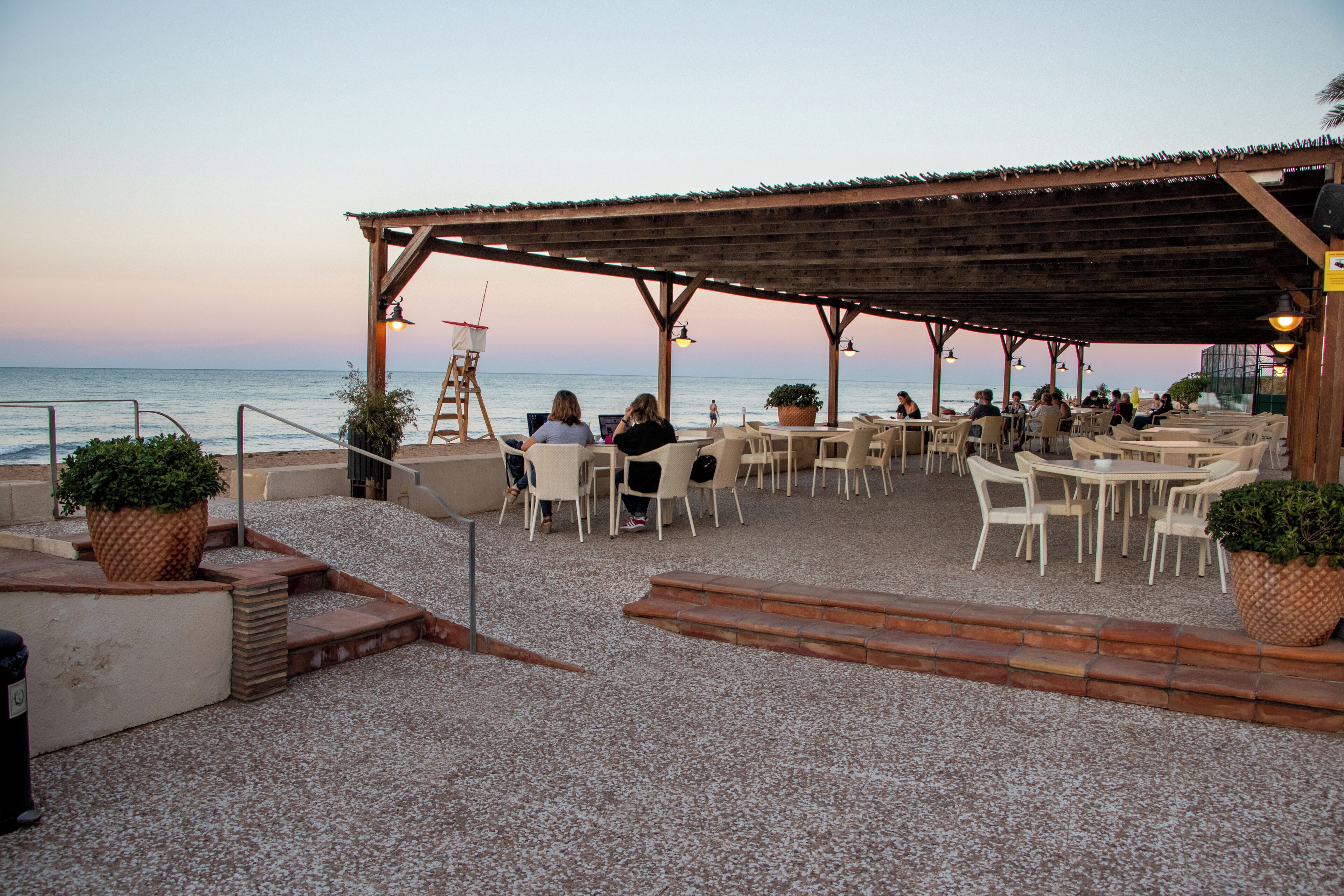 An outdoor seating area with tables and chairs, a wooden pergola, and a view of the sea.