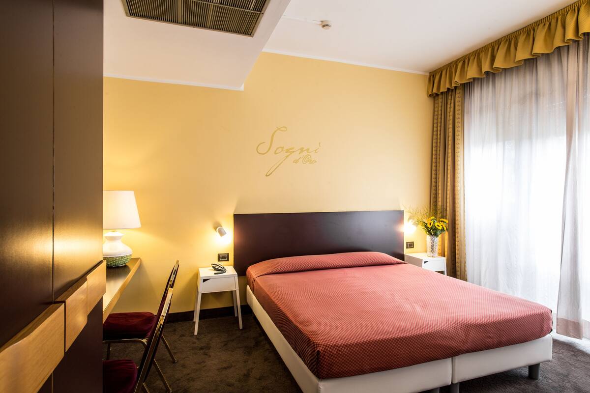 Habitación de hotel con cama, mesita de noche, lámpara, silla y pared con un cartel con el nombre.