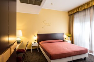 Una camera d'albergo con un letto, un comodino, una lampada, una sedia e un cartello con il nome sulla parete.