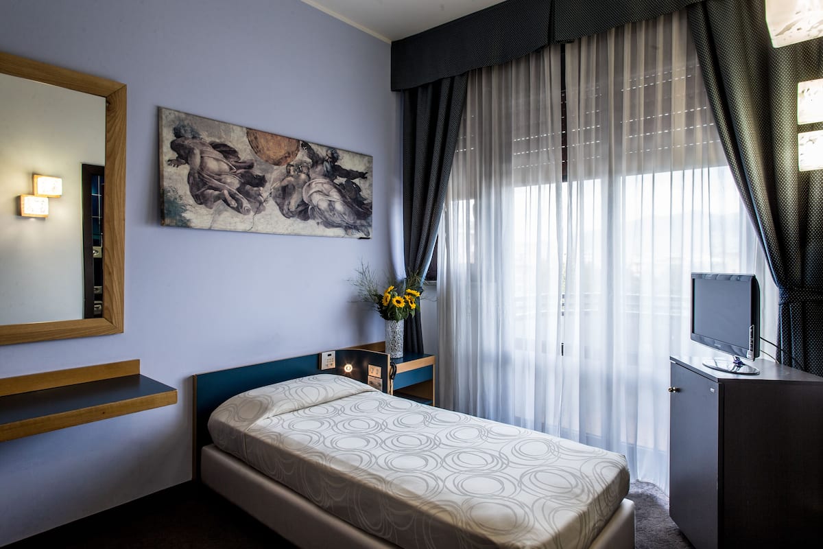 Un dormitorio con cama, escritorio, televisor, un cuadro y ventana con cortinas.
