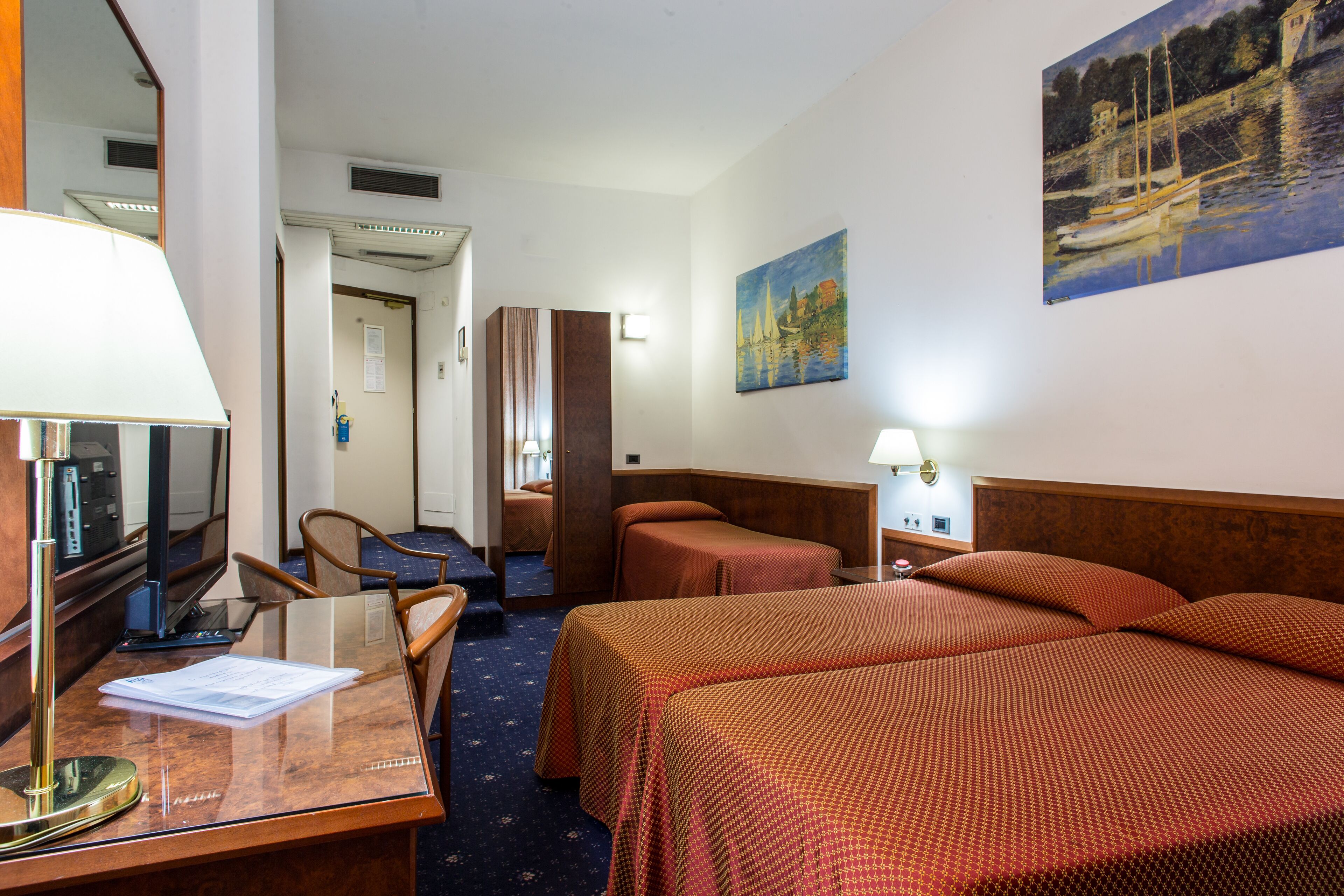Camera d'albergo con due letti, una scrivania con lampada, una sedia, una TV e un quadro appeso al muro.