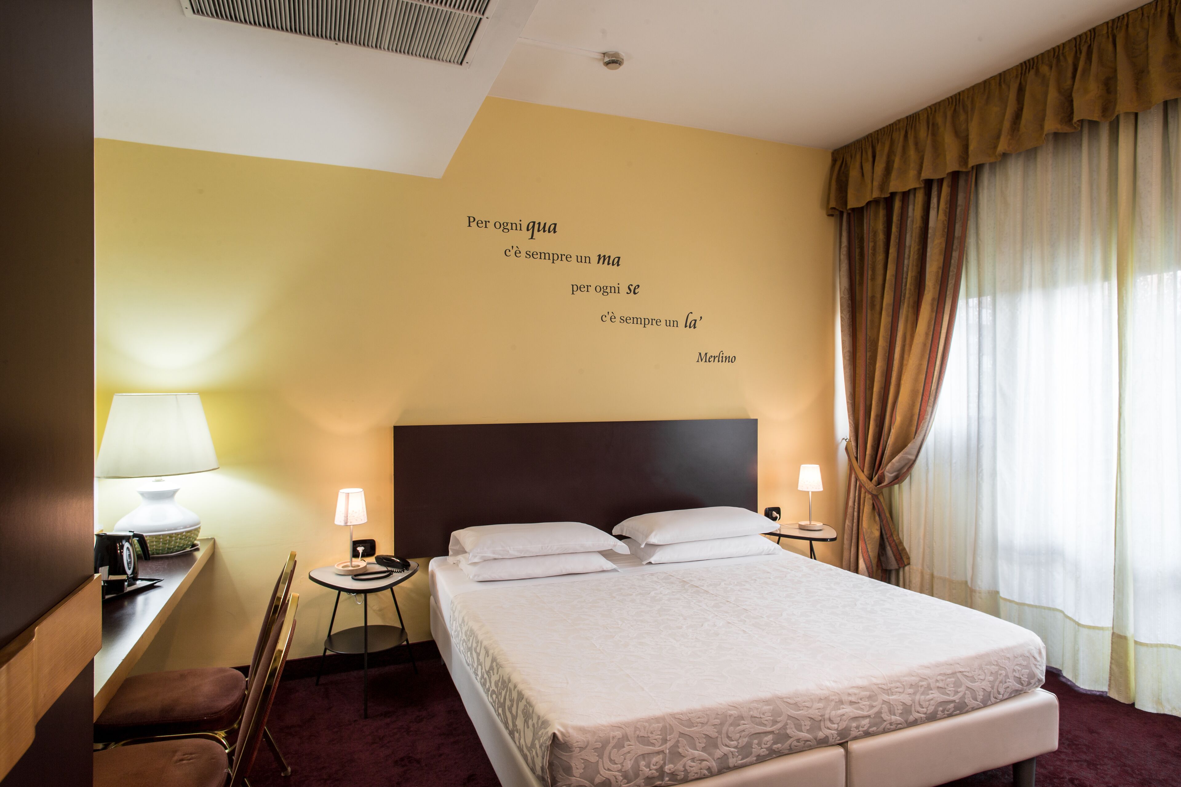 Une chambre d’hôtel avec un grand lit, deux tables de chevet avec des lampes, un bureau avec une chaise et un mur orné d’une citation.