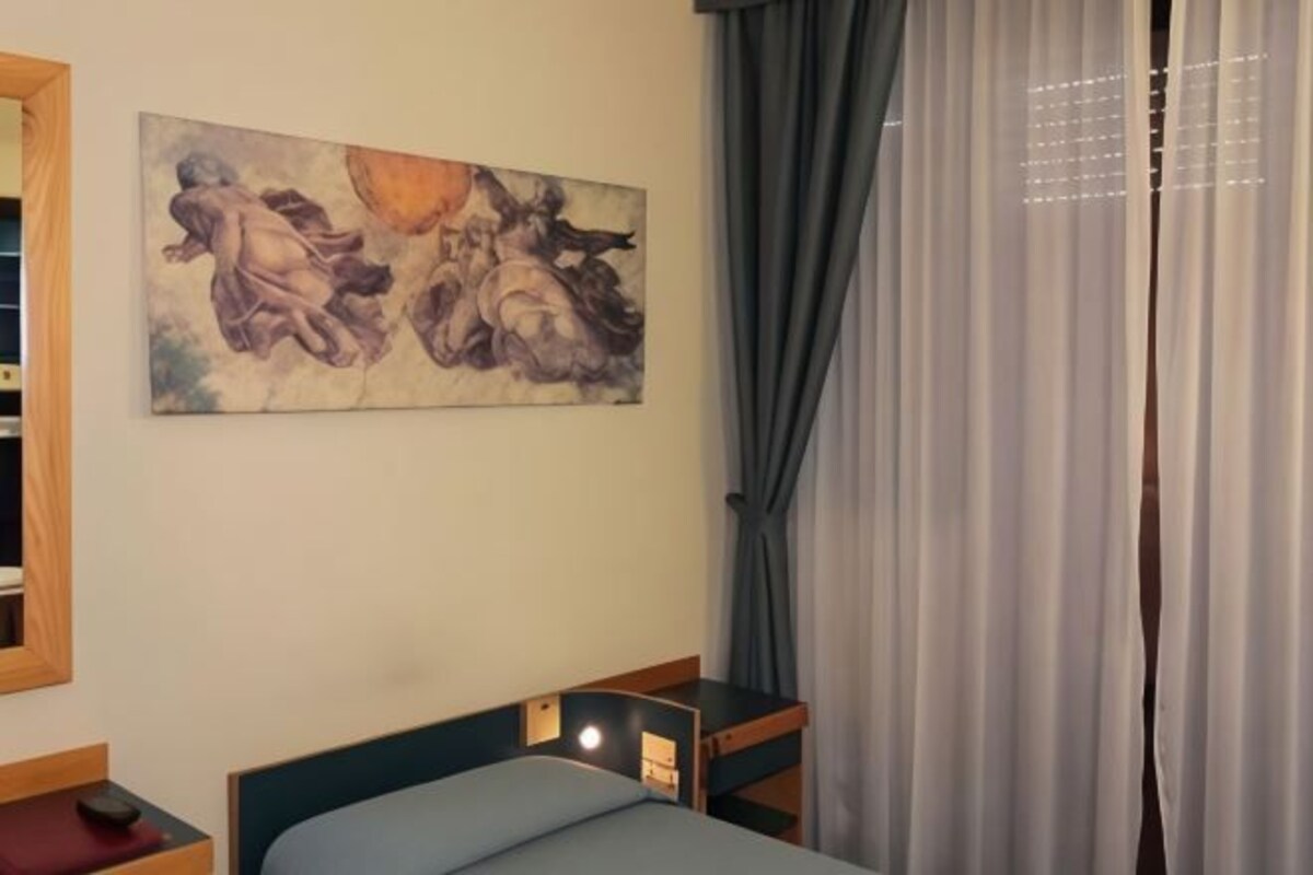 A bedroom with a bed, a nightstand, a painting on the wall, and a window with curtains.