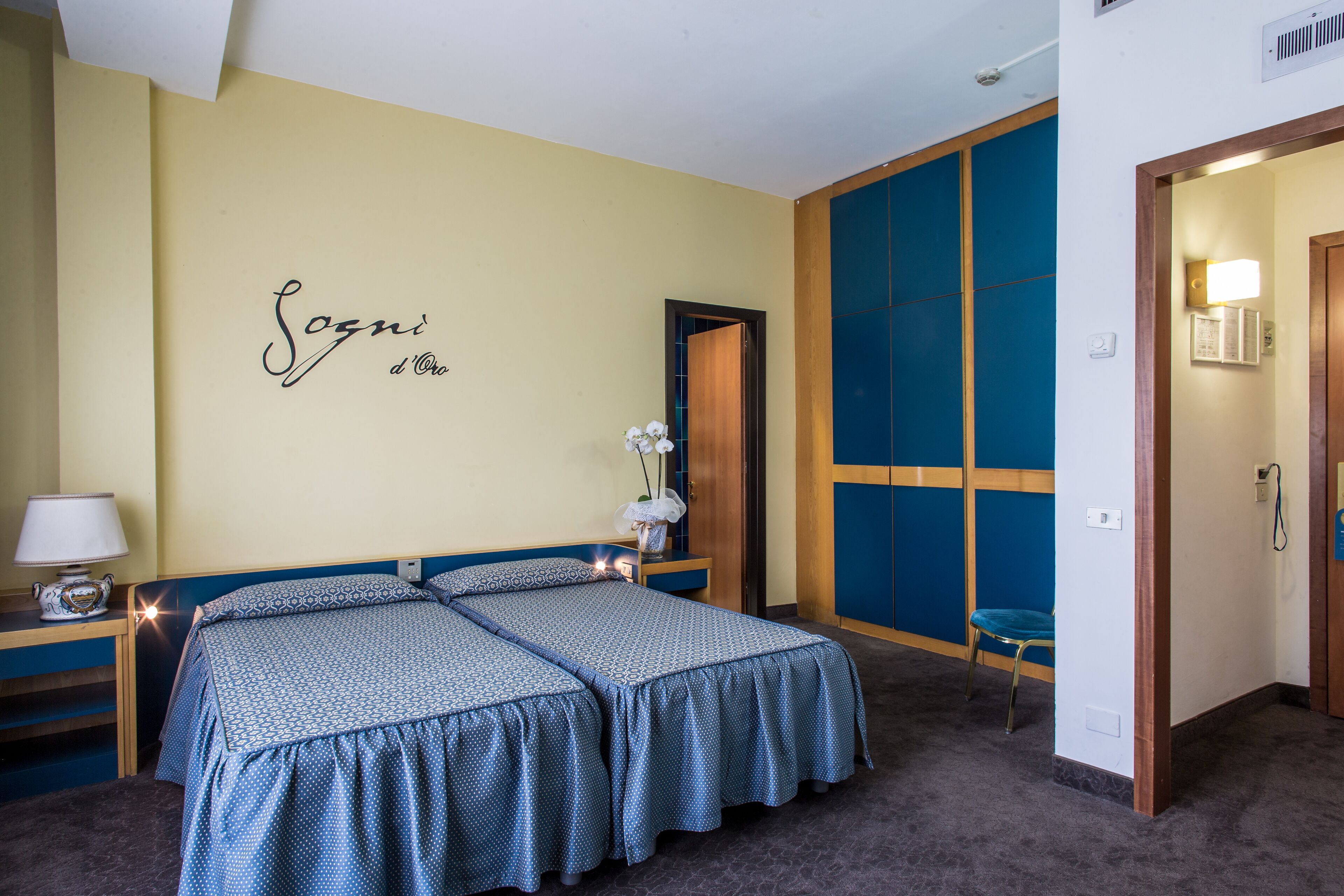 Une chambre d’hôtel avec deux lits, une table de chevet avec une lampe, un élément décoratif mural et une porte donnant sur une autre pièce.