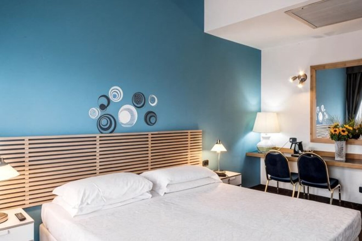 A hotel room with a bed, bedside tables, a desk with chairs, a lamp, and a wall decoration.