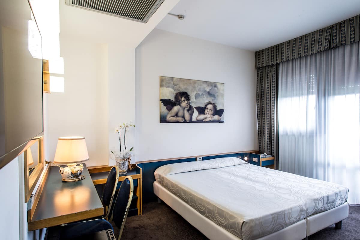 Habitación de hotel con cama, escritorio con lámpara, una silla, un cuadro en la pared y ventana con cortinas.