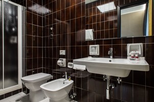 Et moderne badeværelse med et hvidt toilet, en bidet, en vask med spejl og mørkebrune fliser.
