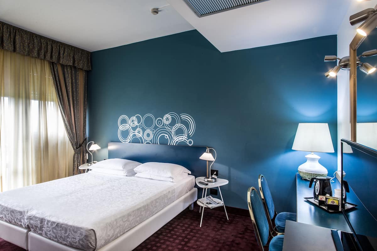 Habitación de hotel con una cama grande, dos mesitas de noche con lámparas, un escritorio con silla, un espejo y un adorno en la pared.
