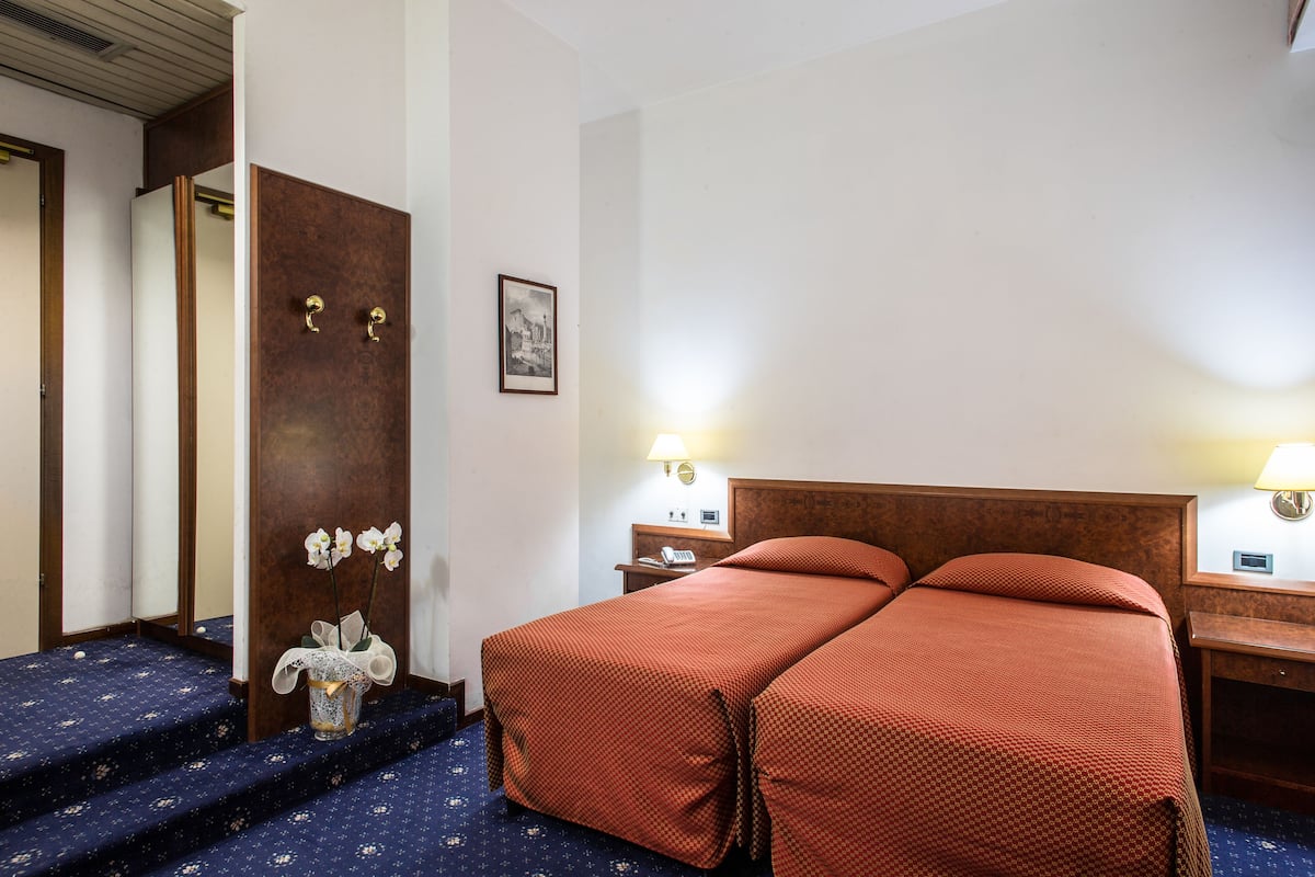 Habitación de hotel con cama, dos mesitas de noche, una lámpara, un cuadro en la pared y un pequeño florero con flores en el suelo.