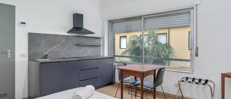 Une chambre d’hôtel moderne équipée d’un lit, d’une petite table, d’une chaise, d’une fenêtre donnant sur des arbres et d’une kitchenette.