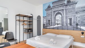 Une chambre d’hôtel moderne équipée d’un lit, d’un bureau en bois, d’une chaise, d’une armoire et ornée d’une fresque représentant un bâtiment historique.