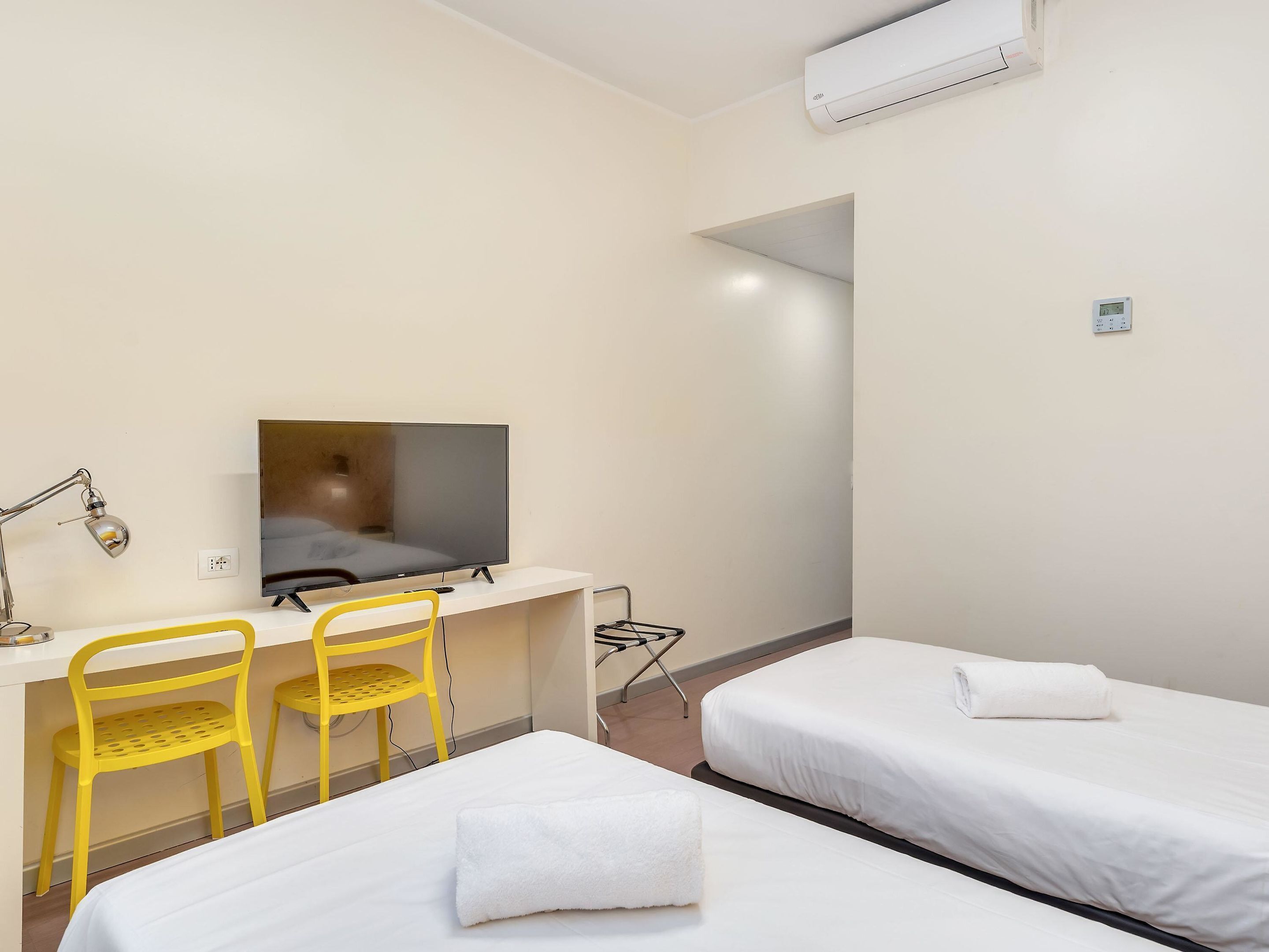 Ett hotellrum med ett skrivbord, två gula stolar, en tv, en säng med en vit handduk och en luftkonditioneringsanläggning.