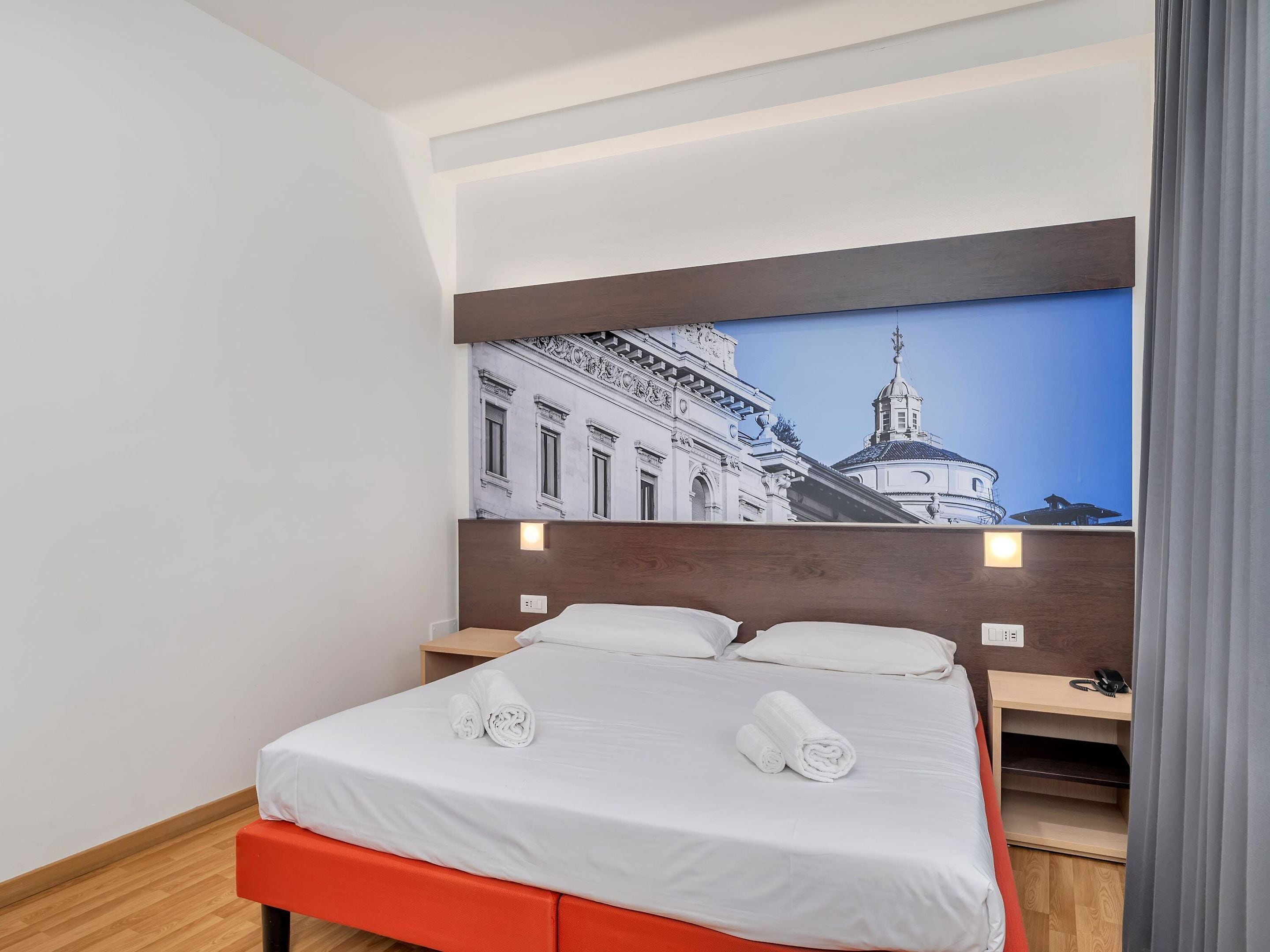 A hotel room with a bed, two nightstands, a large wall-mounted picture of a building, and a window with curtains.