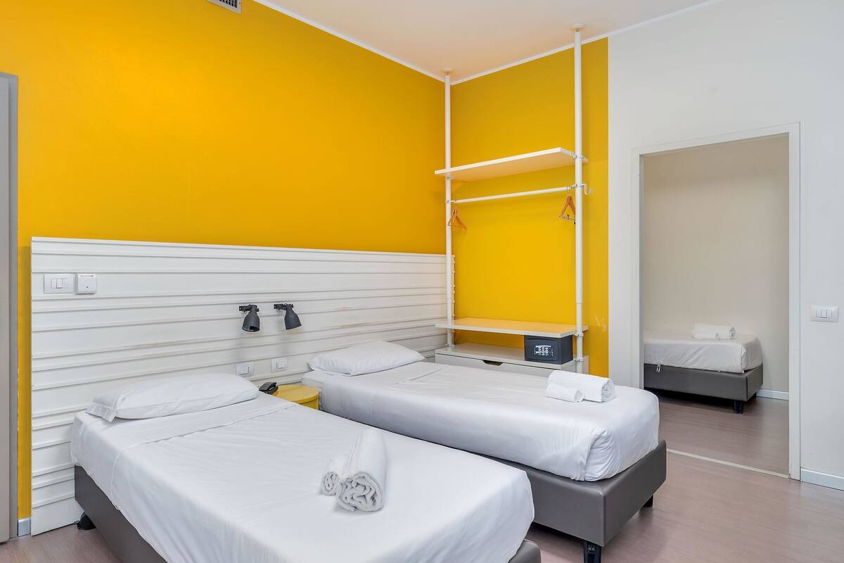 Hotellihuone, jossa on kaksi sänkyä, valkoinen pääty, yksi keltainen seinä, pieni yöpöytä lampun kanssa ja vaatekaappi.