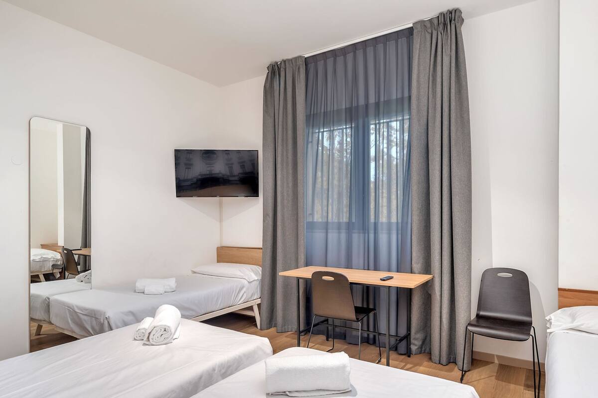 Hotellihuone, jossa on kaksi sänkyä, työpöytä, tuoli, televisio ja suuri ikkuna, jossa on verhot.