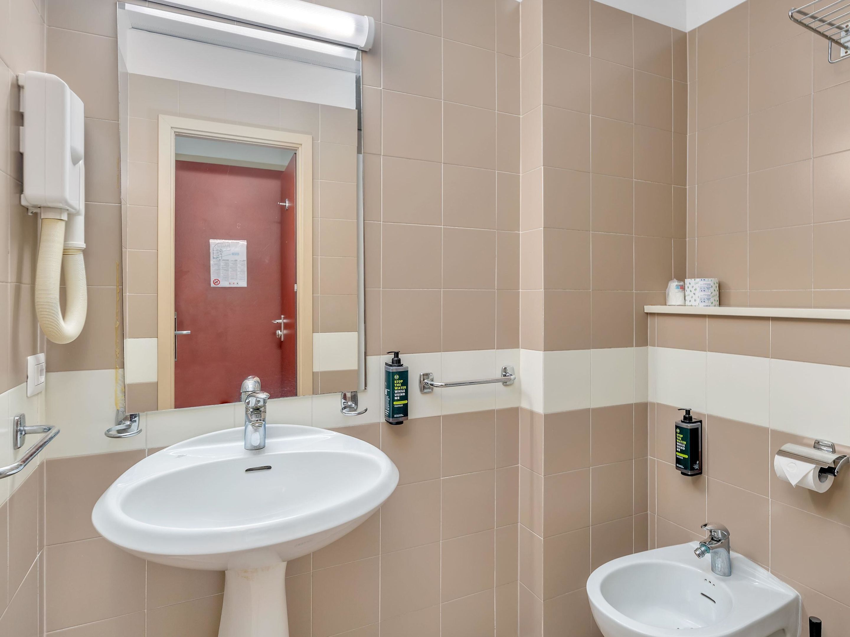 Une salle de bain avec un lavabo blanc, un petit lavabo, un miroir, un porte-serviettes et un distributeur de papier toilette.
