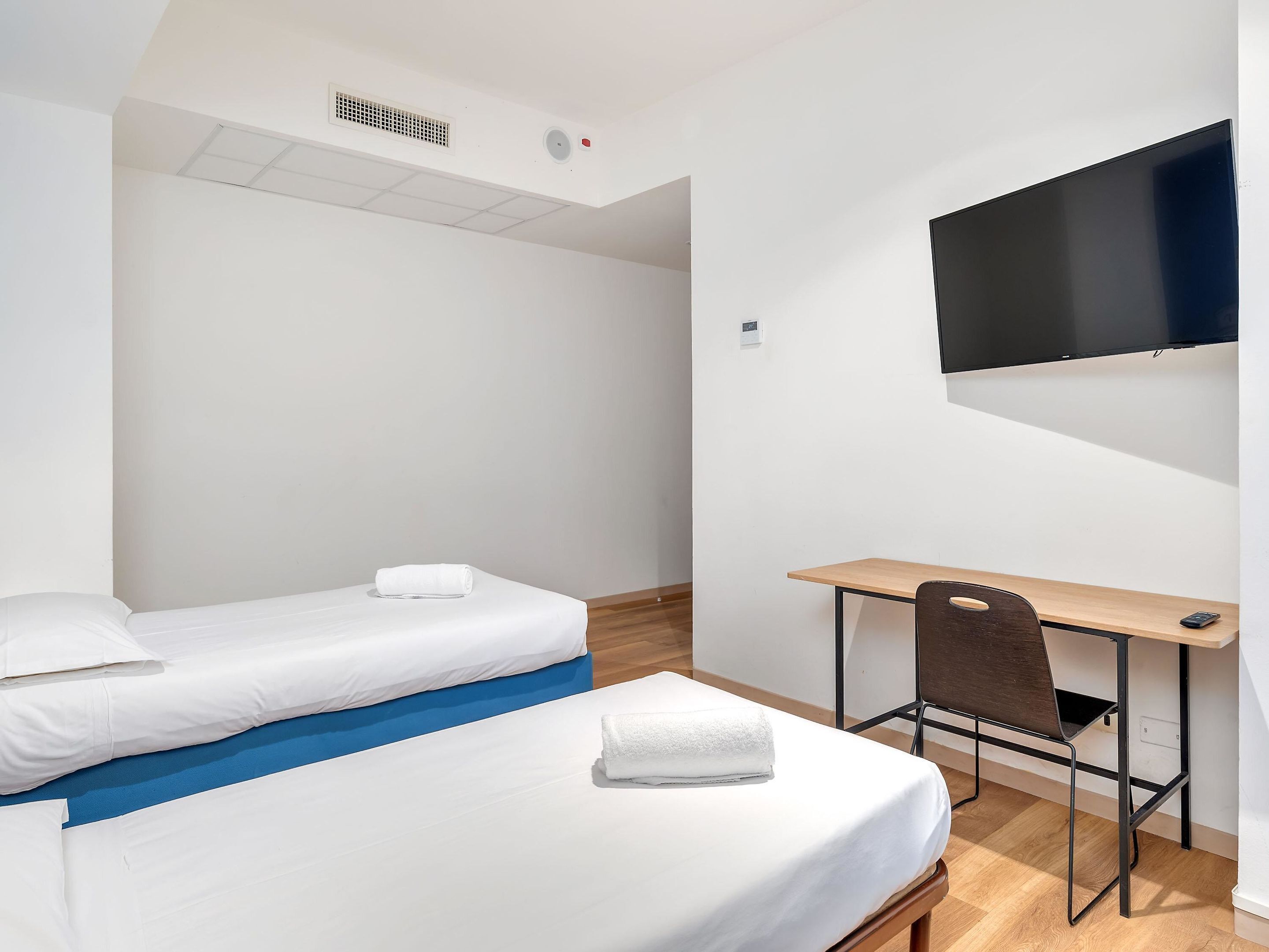 Une chambre d’hôtel avec deux lits simples, un bureau, une chaise, un téléviseur à écran plat fixé au mur et une bouche d’aération au plafond.