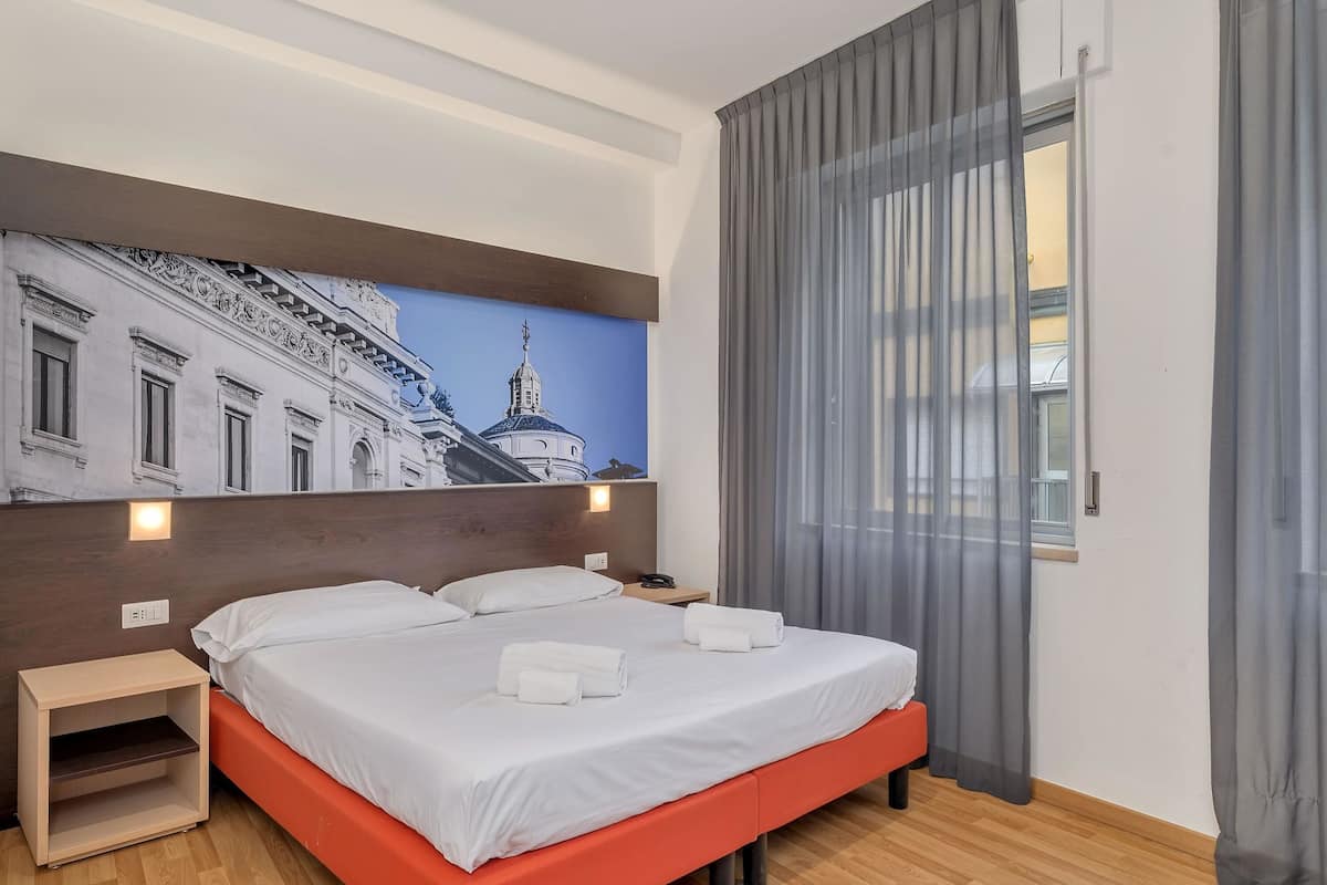 Hotellihuone, jossa on sänky, yöpöytä, seinään kiinnitetty valaisin, ikkuna verhoilla ja seinämaalaus, jossa on kuvattuna kaupunkimaisema.