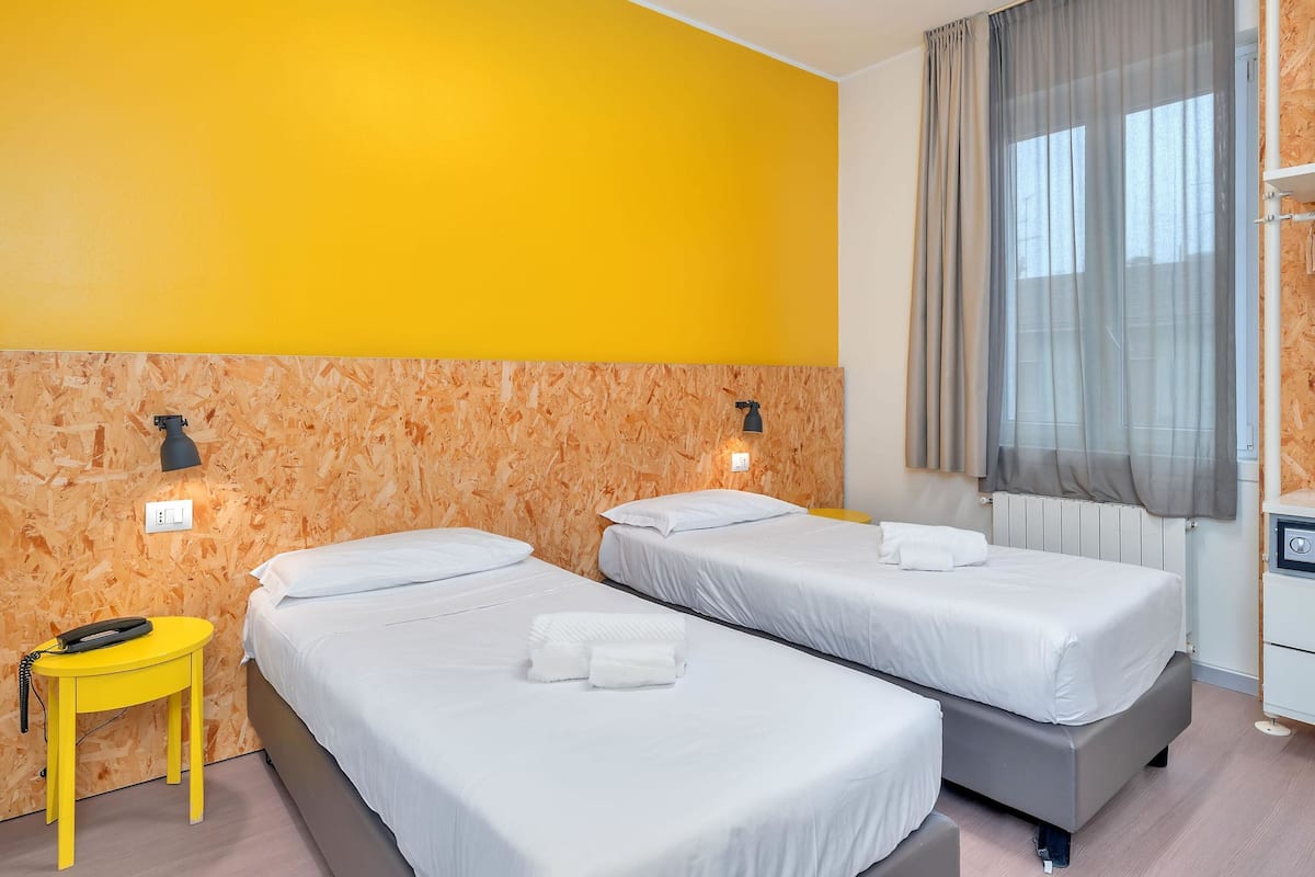 Hotellihuone, jossa on kaksi sänkyä, puinen pääty, keltainen yöpöytä ja ikkuna, jossa on verhot.